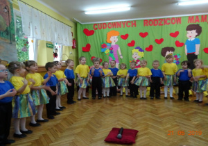 Dziewczynki ubrane w żółte podkoszulki i zielono-żółtto-niebieskie spódniczki oraz chłopcy w niebieskich podkoszulkach i czarnych spodniach śpiewają piosenkę dla rodziców.
